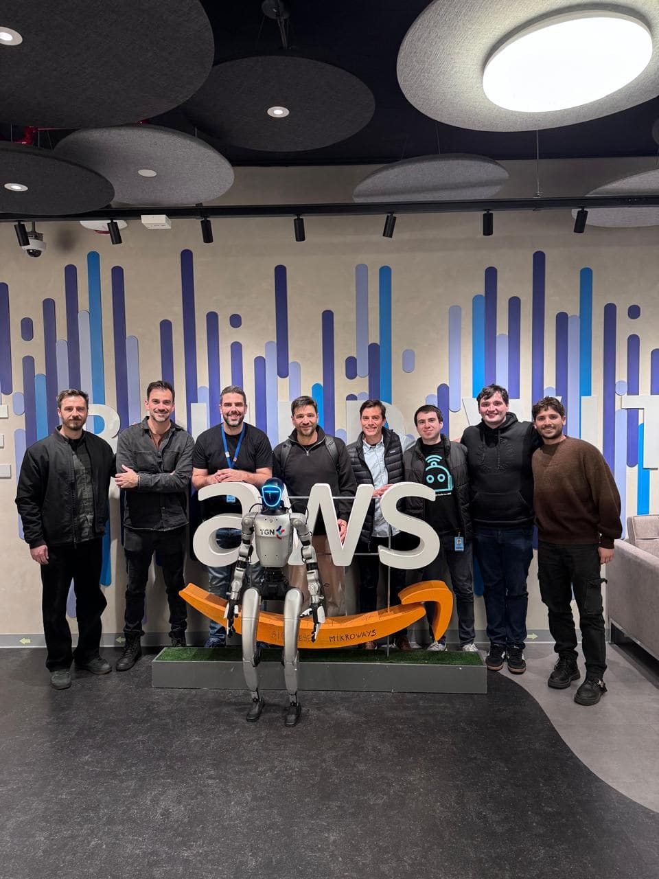 Eight people lined up behind an AWS logo installation, a small robot standing on the plinth in front of them.