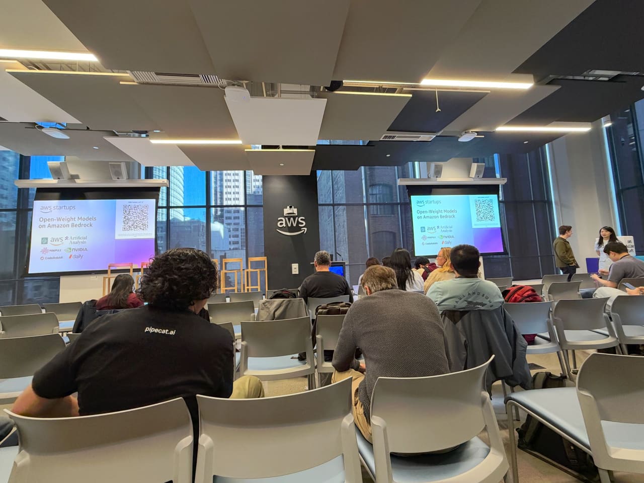 An audience at the AWS Builder Loft facing twin screens announcing a session on open-weight models on Amazon Bedrock.