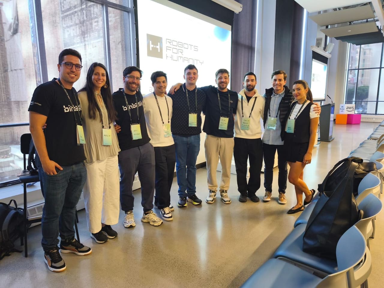 The binbash and Robots for Humanity teams together in front of the Robots for Humanity title slide, at the AWS Builder Loft in San Francisco.